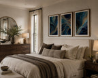 Close-up view of the Blue Gold Modern Abstract Triptych Printable Wall Art Set of 3 displayed above a walnut dresser in a bright contemporary bedroom. Blue and gold abstract three-panel gallery wall prints in natural oak frames with elegant white matting pair beautifully with designer décor, candles, books, soft bedding, and warm afternoon sunlight for a luxury interior aesthetic.