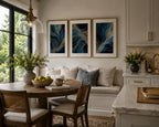 Blue Gold Modern Abstract Triptych Printable Wall Art Set of 3 hanging above a built-in breakfast bench in a bright farmhouse kitchen. Three blue and gold abstract printable gallery wall prints in refined wood frames with white matting complement marble countertops, brass hardware, round wooden dining table, black framed windows, and fresh floral arrangements. Instant download wall art for modern kitchens.