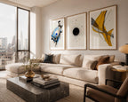 Sophisticated New York apartment bedroom featuring an oversized contemporary watercolor abstract gallery wall in expressive shades of cobalt blue, charcoal black and warm mustard yellow. The elegant triptych is professionally framed in natural wood with oversized white mat boards above the upholstered headboard, while angled afternoon sunlight creates subtle reflections that enhance the luxurious urban interior.