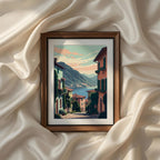 Cobblestone street descending through a colorful Italian hillside village toward a peaceful Mediterranean lake surrounded by mountains under a pastel sunset sky. Vintage Italian coastal landscape printable wall art, framed artwork, canvas print, poster and digital home decor.